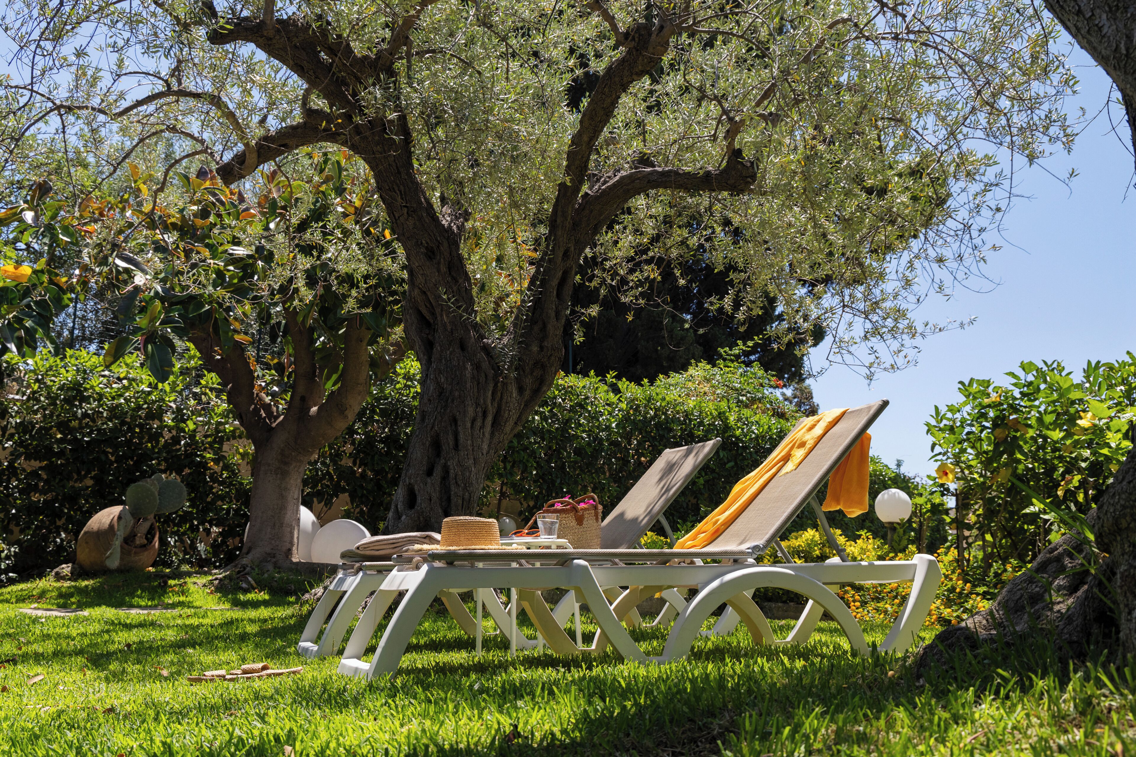 A garden with a large tree, a white lounge chair, and a sun umbrella.