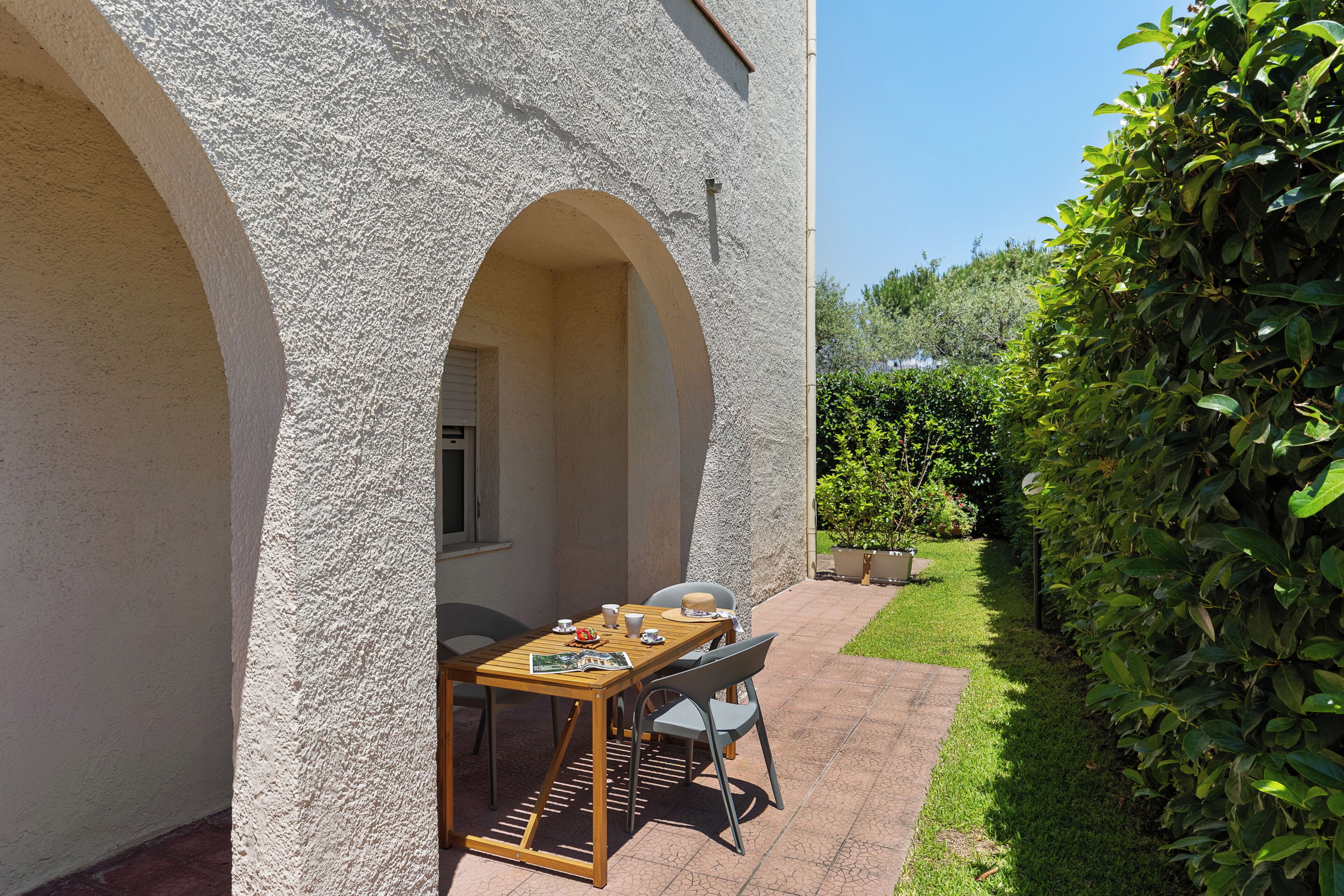 Eine Terrasse mit Tisch und Stühlen, eine weiße Wand mit strukturierter Oberfläche und ein grüner Heckenstreifen.
