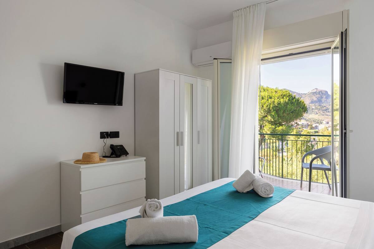 A hotel room with a bed, a TV mounted on the wall, a wardrobe, a desk with a phone and a hat, and a balcony with a view of trees and mountains.