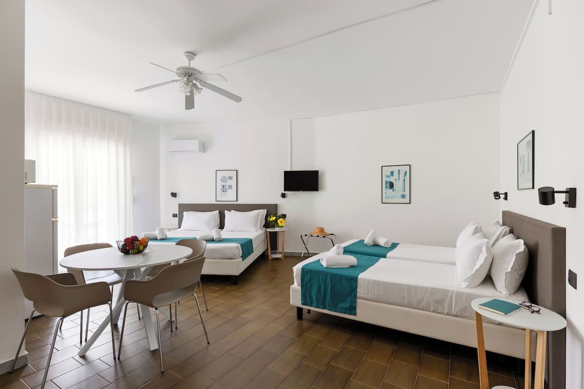 A hotel room with two beds, a dining table, chairs, a ceiling fan, a TV, and artwork on the walls.