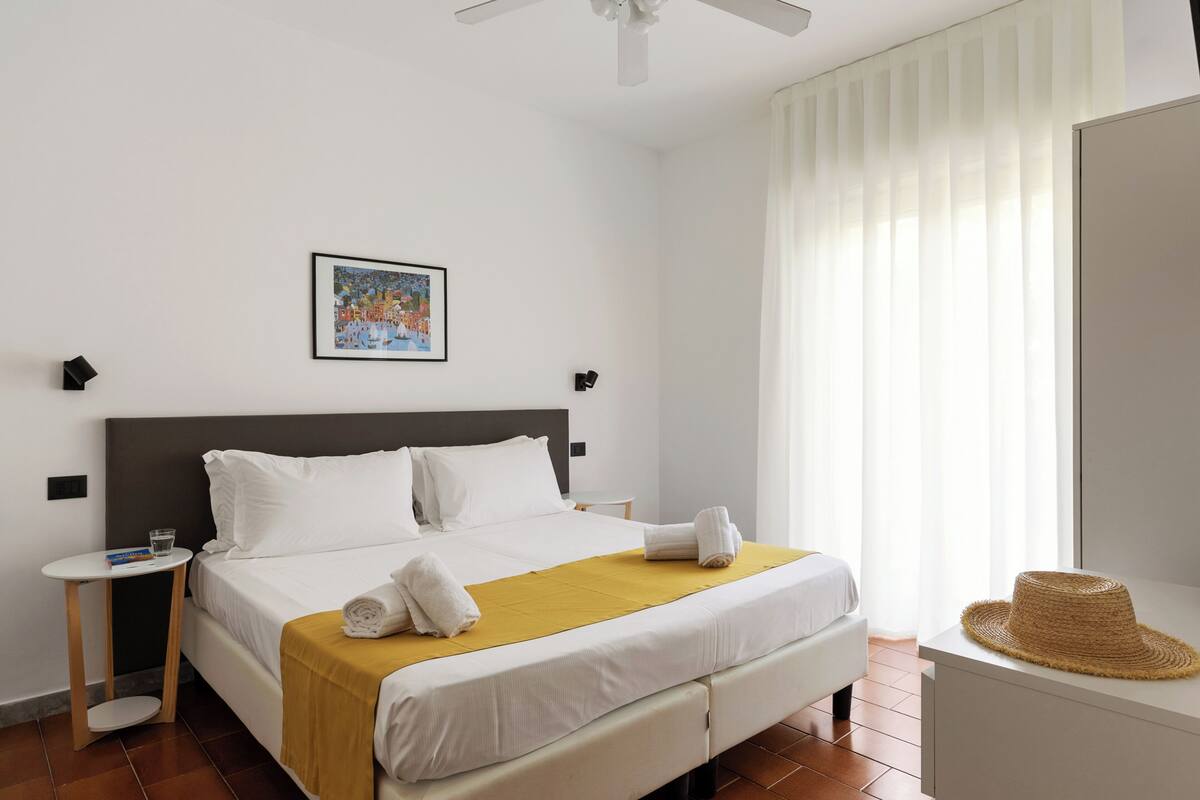 A hotel room with a bed, a bedside table, a wall-mounted picture, a ceiling fan, and a window with curtains.