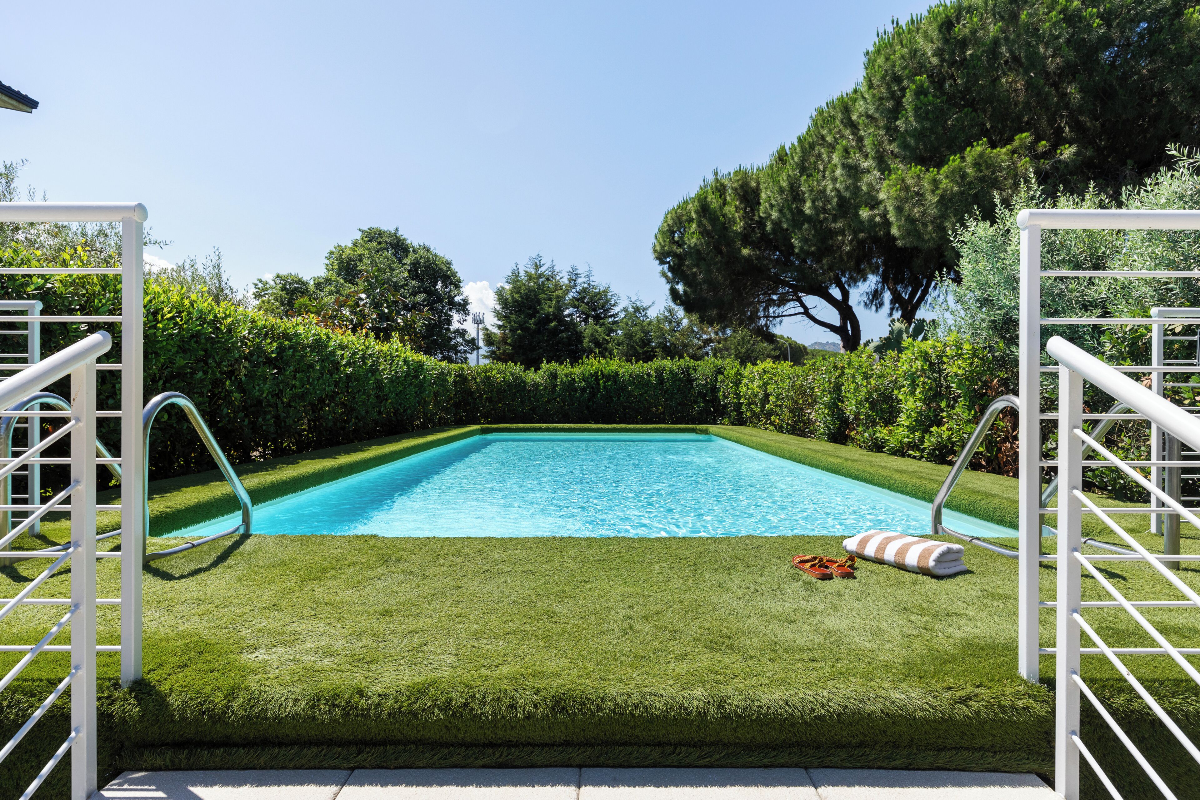 Une piscine rectangulaire dotée d’une plage blanche et de garde-corps en métal, entourée de pelouse et de haies parfaitement entretenues.