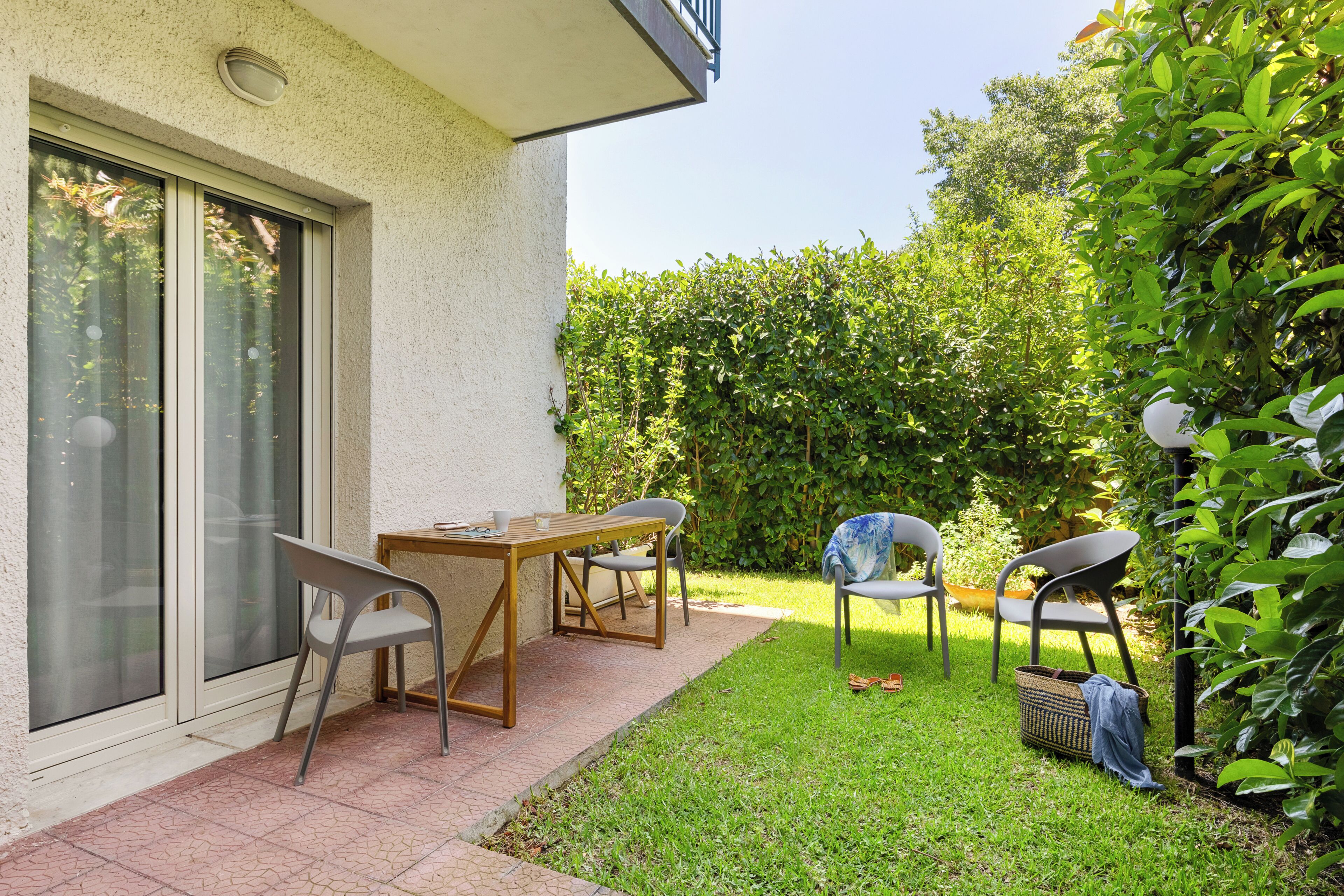 Une terrasse avec une table et des chaises, une fenêtre avec des stores et une haie en arrière-plan.