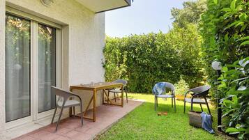 Une terrasse avec une table et des chaises, une fenêtre avec des stores et une haie en arrière-plan.