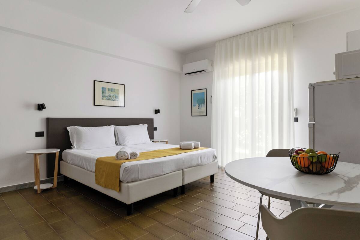 A hotel room with a bed, bedside table, a round dining table with chairs, and a fruit bowl.