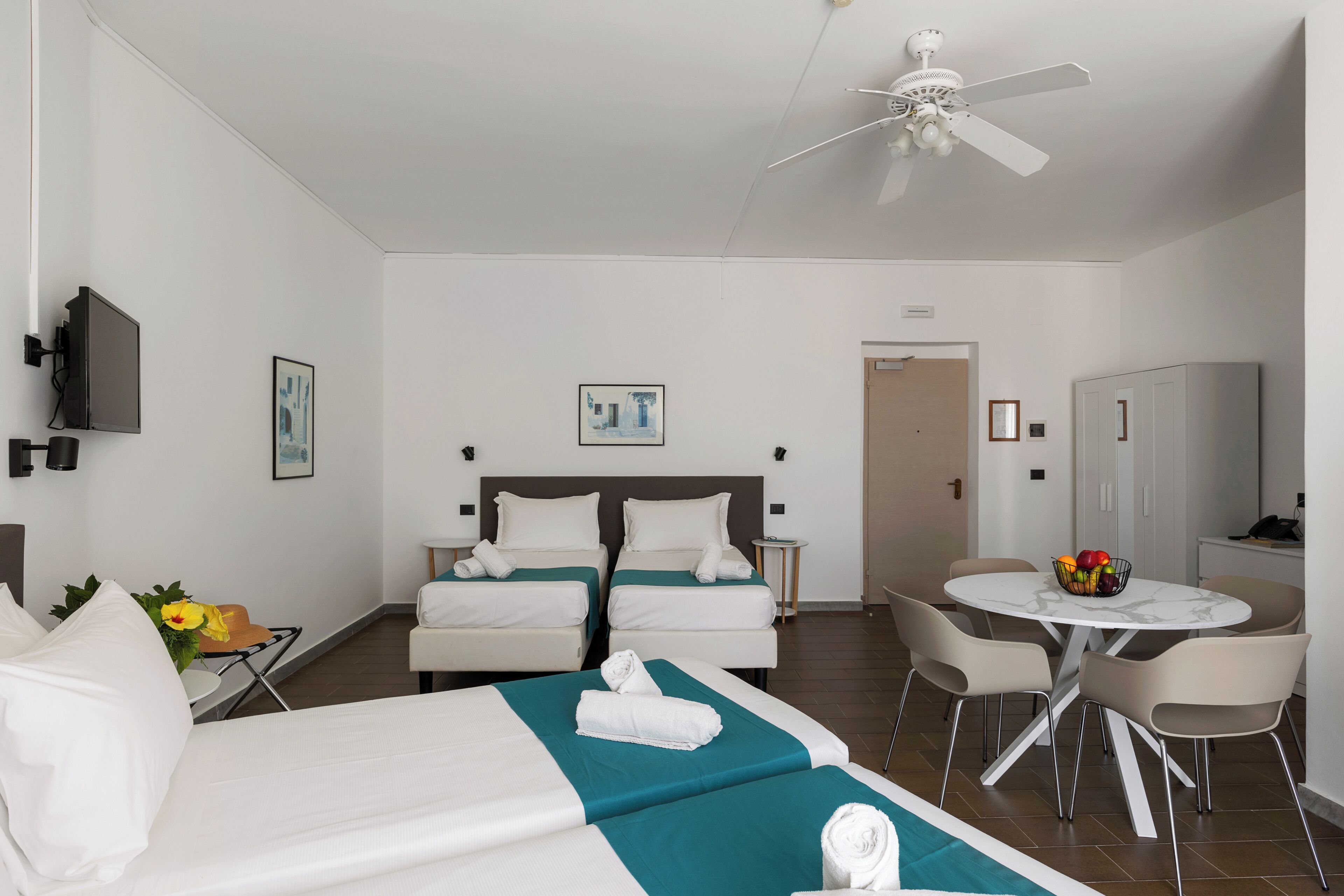 Ein Hotelzimmer mit zwei Betten, einem Esstisch mit Stühlen, einem Deckenventilator und einem kleinen Fernseher an der Wand.