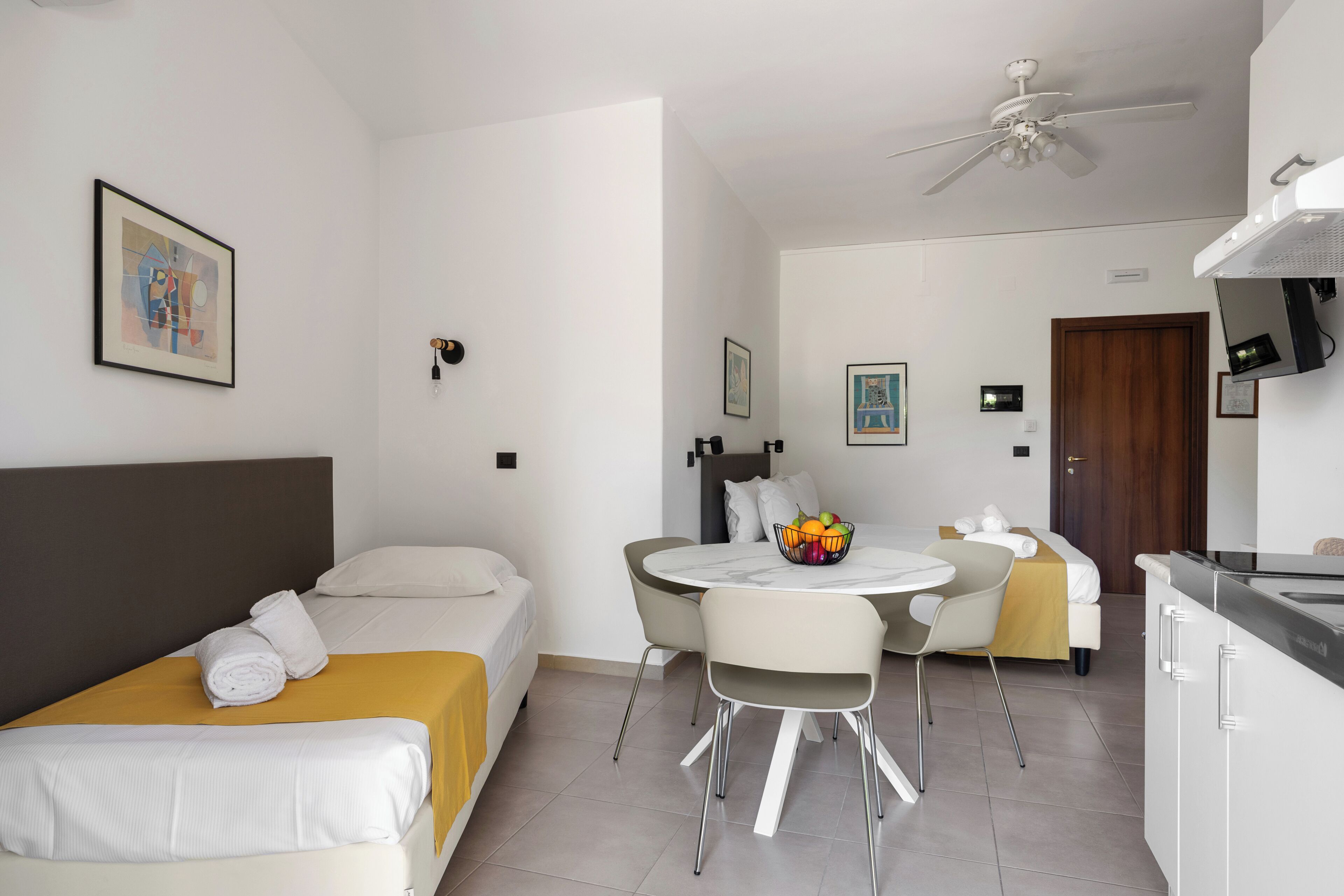 A hotel room with a bed, a dining table with chairs, a kitchen area, and a ceiling fan.