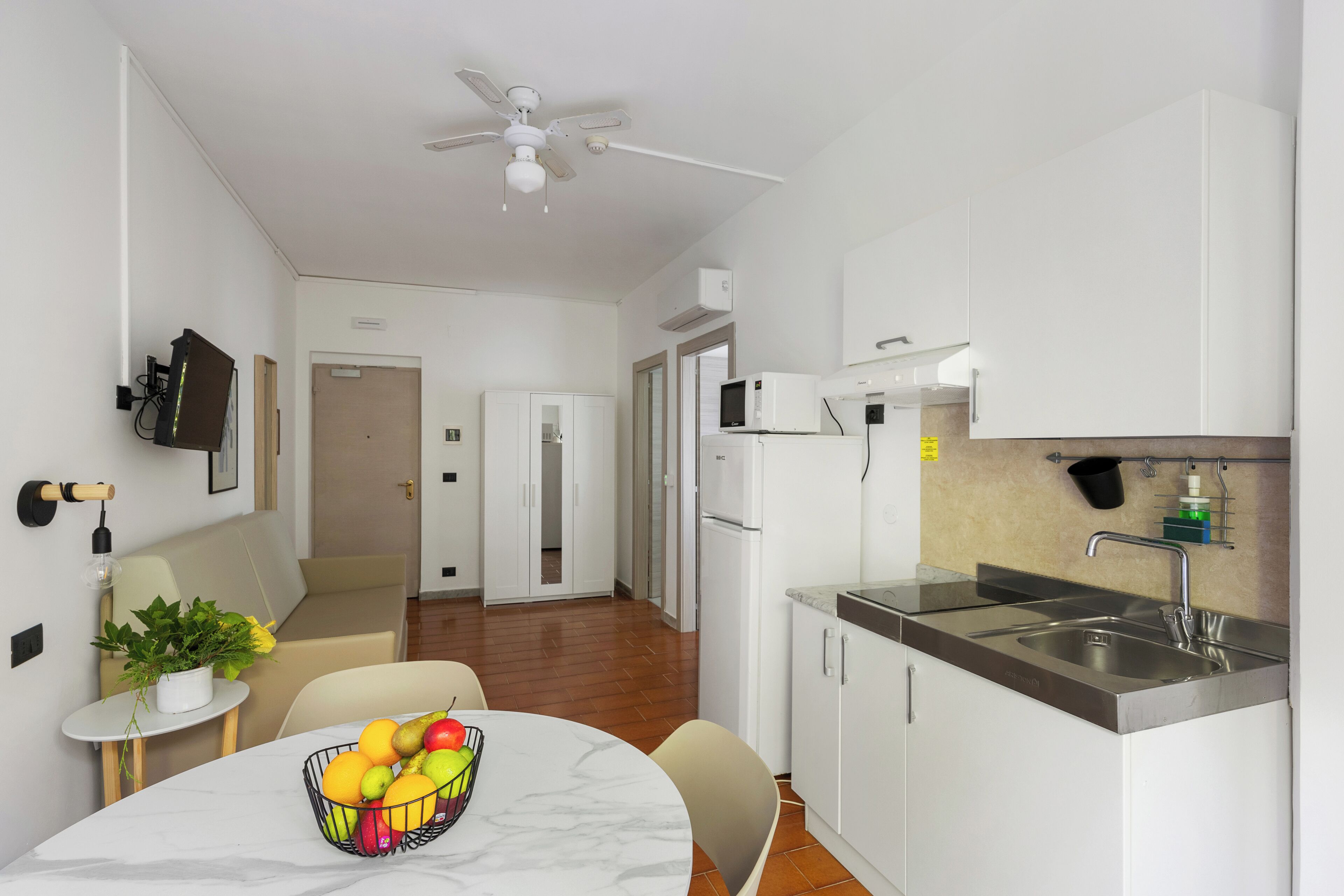 A modern kitchen with white cabinets, a stainless steel sink, and a dining area with a marble table and a fruit bowl.