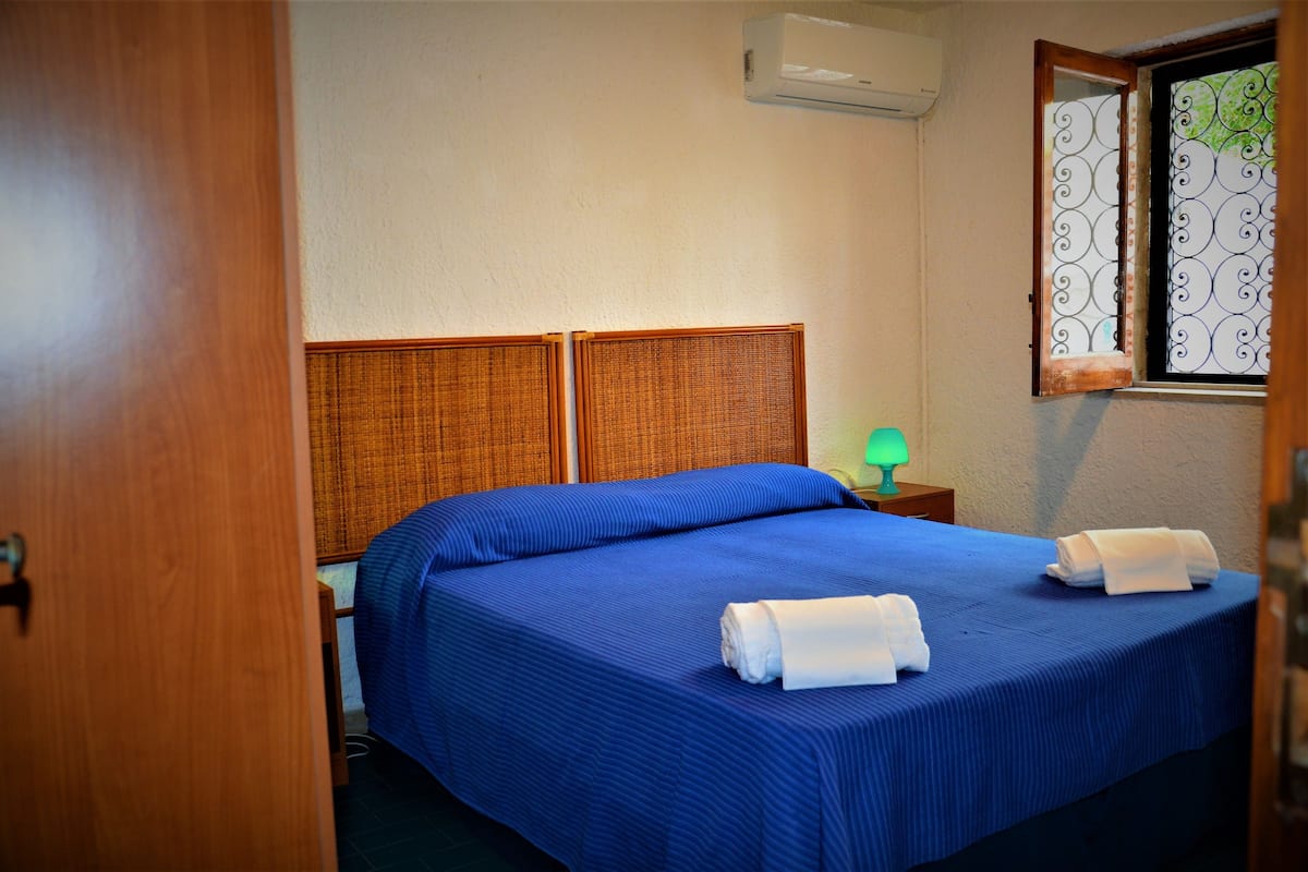 Ett hotellrum med en sänggavel i trä, ett blått överkast, två hoprullade handdukar, en grön lampa och ett fönster med dekorativa järngaller.