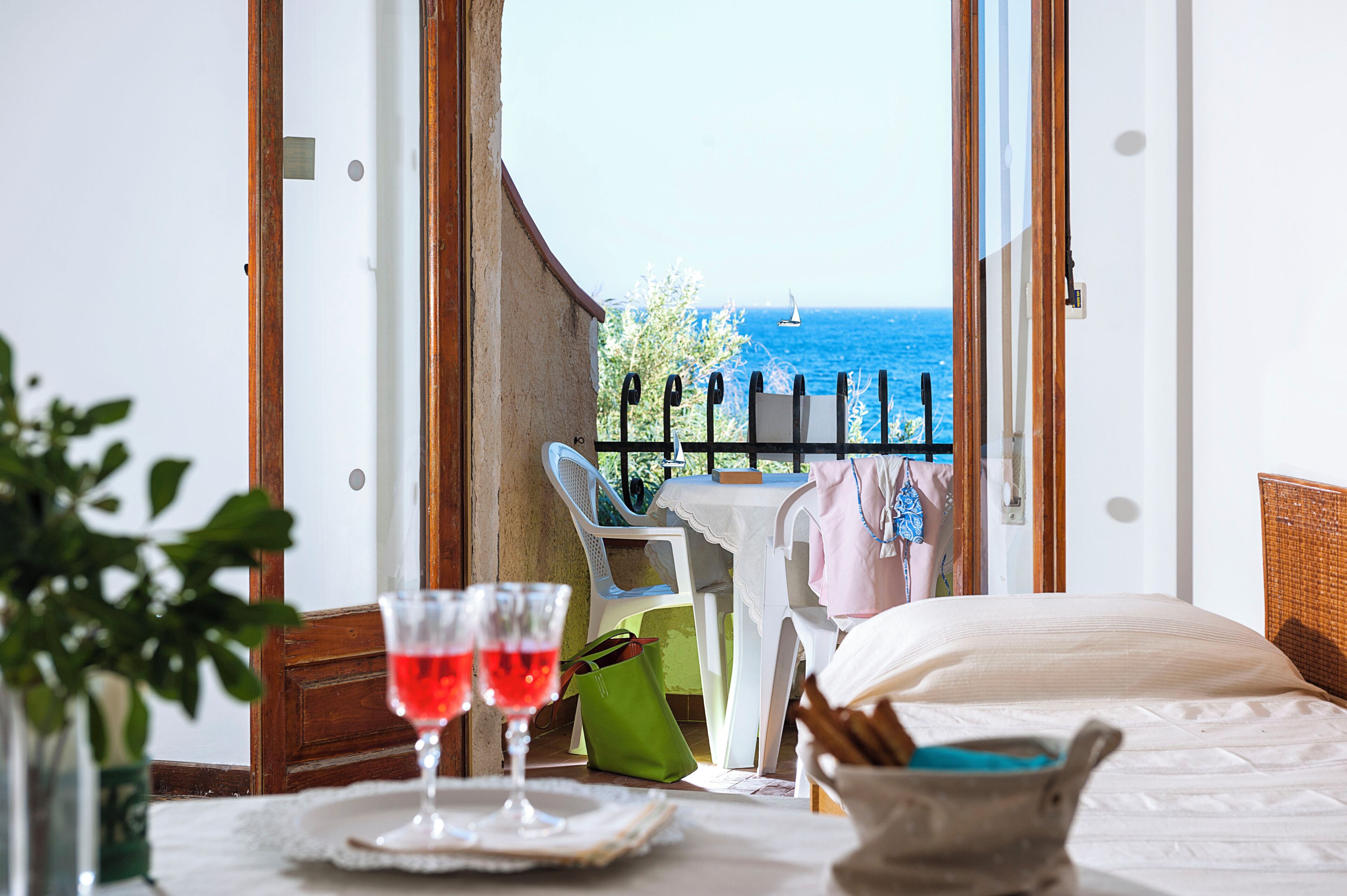 Une chambre d’hôtel avec un balcon, un lit, une table avec deux verres de vin rouge et une vue sur la mer.