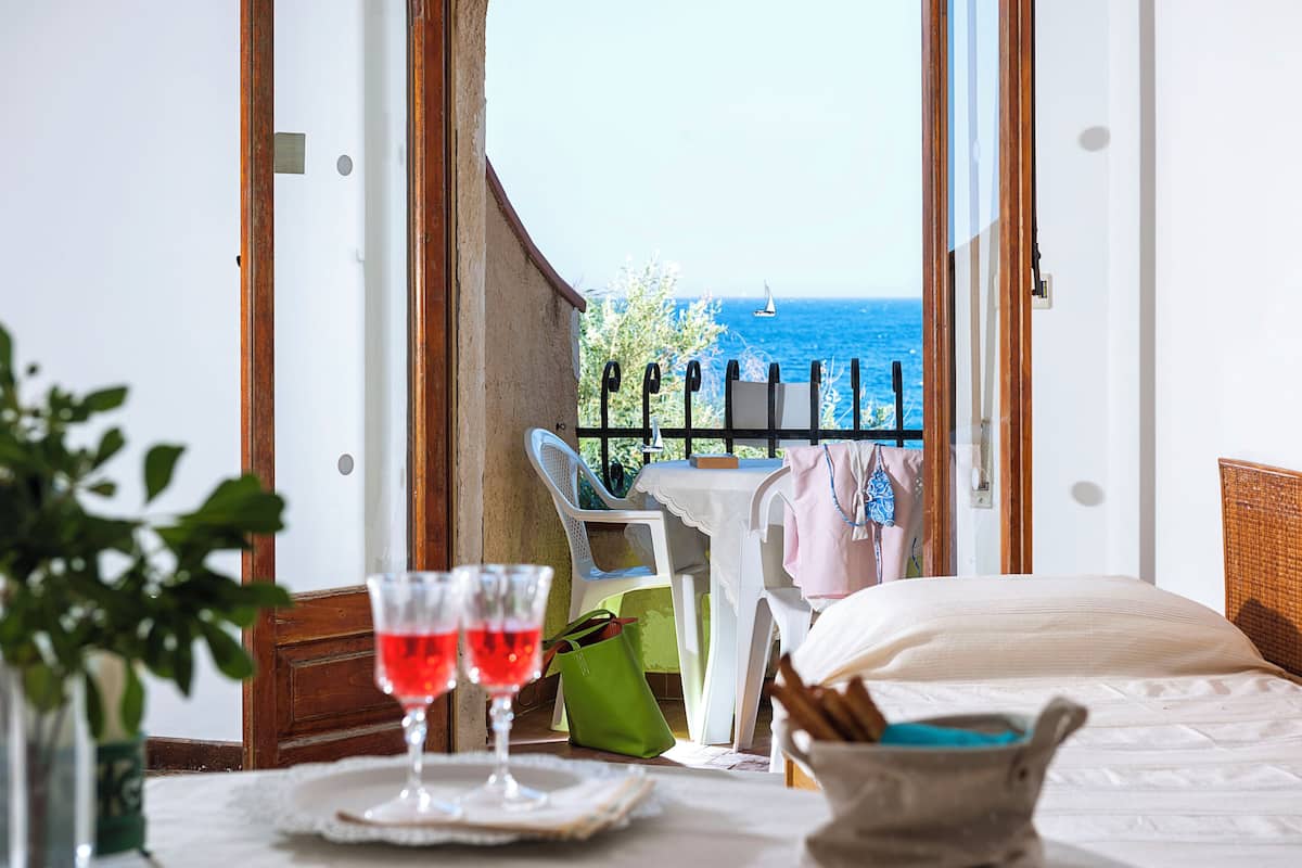 Une chambre d’hôtel avec un balcon, un lit, une table avec deux verres de vin rouge et une vue sur la mer.