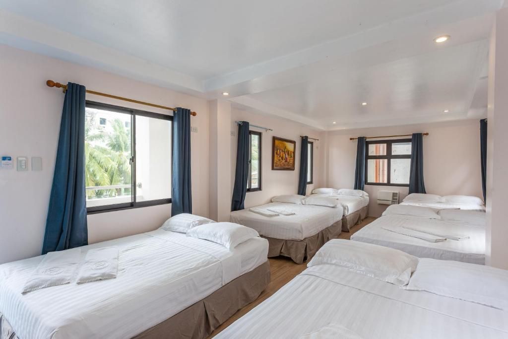 A hotel room with four single beds, a large window with curtains, a painting on the wall, and a ceiling with recessed lighting.