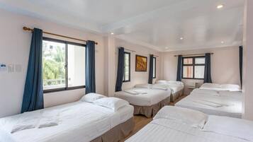 A hotel room with four single beds, a large window with curtains, a painting on the wall, and a ceiling with recessed lighting.