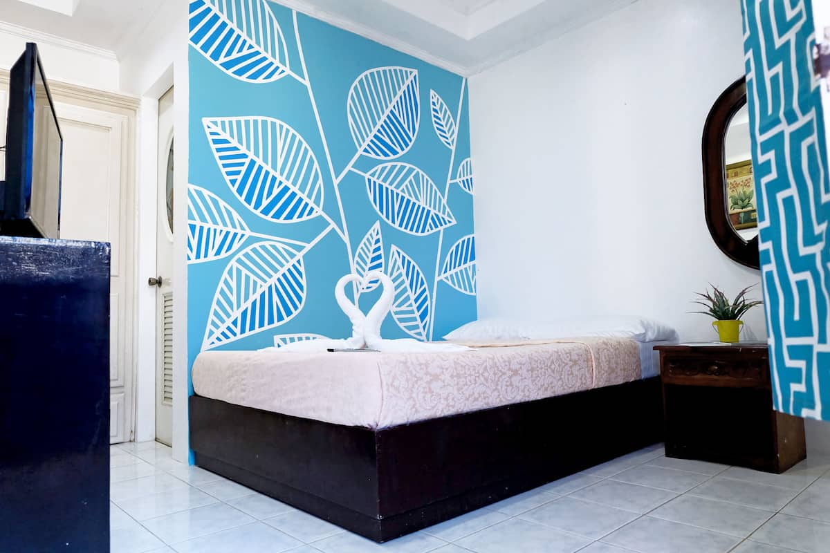 A bedroom with a bed, a wall with a leaf pattern, a small table with a plant, and a mirror on the wall.