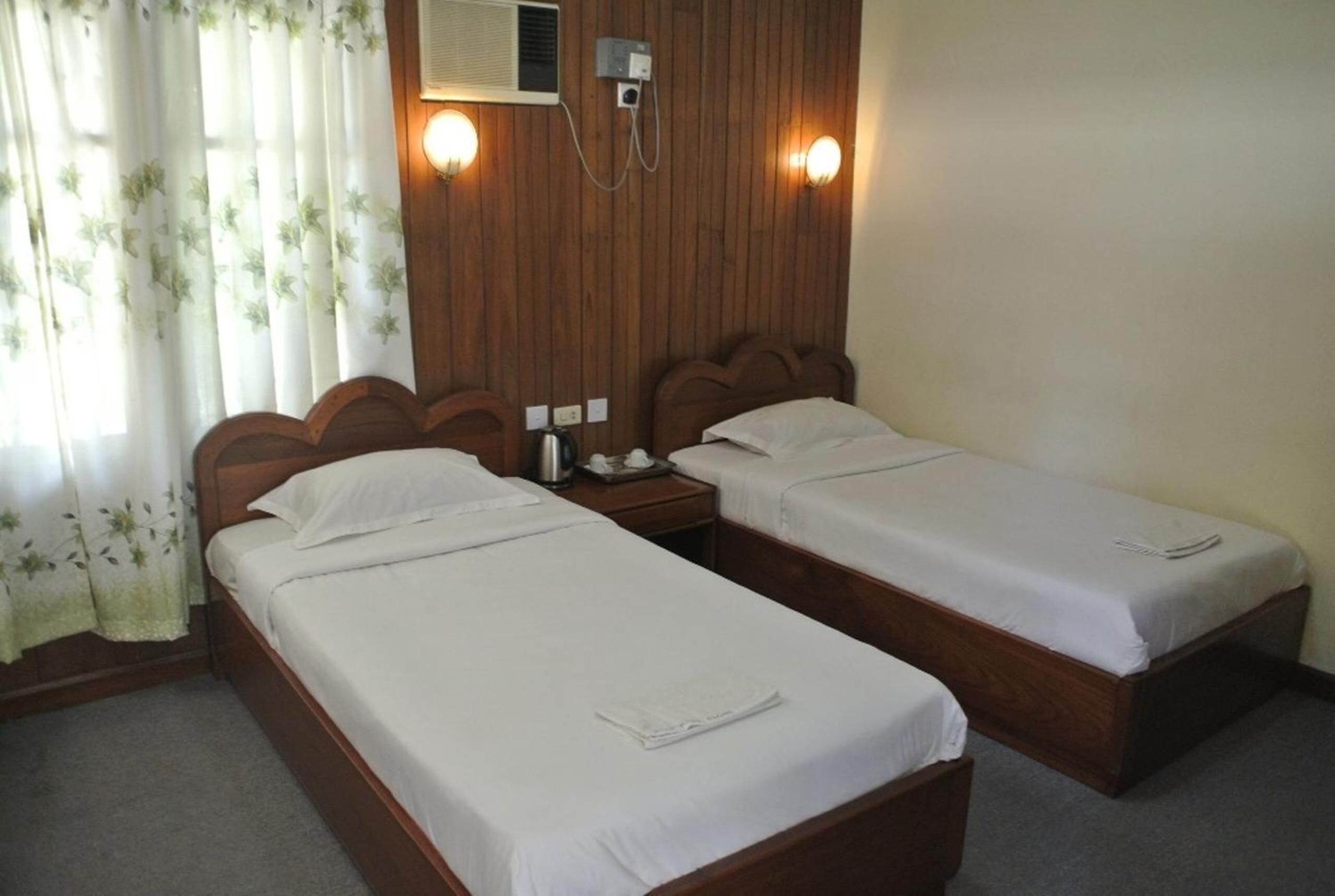 A hotel room with two single beds, a wooden headboard, a bedside table with a lamp, and a wall-mounted air conditioner.