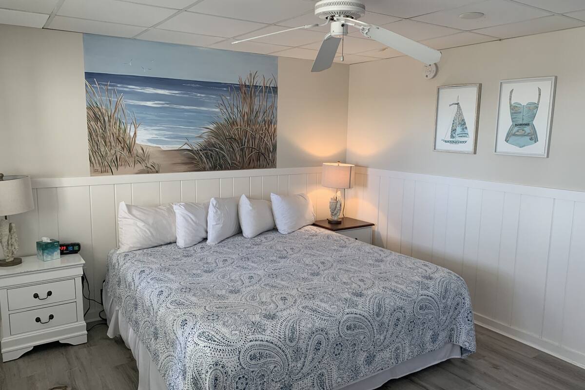 A bedroom with a bed, nightstands, a ceiling fan, a painting of a beach, and framed artwork on the wall.