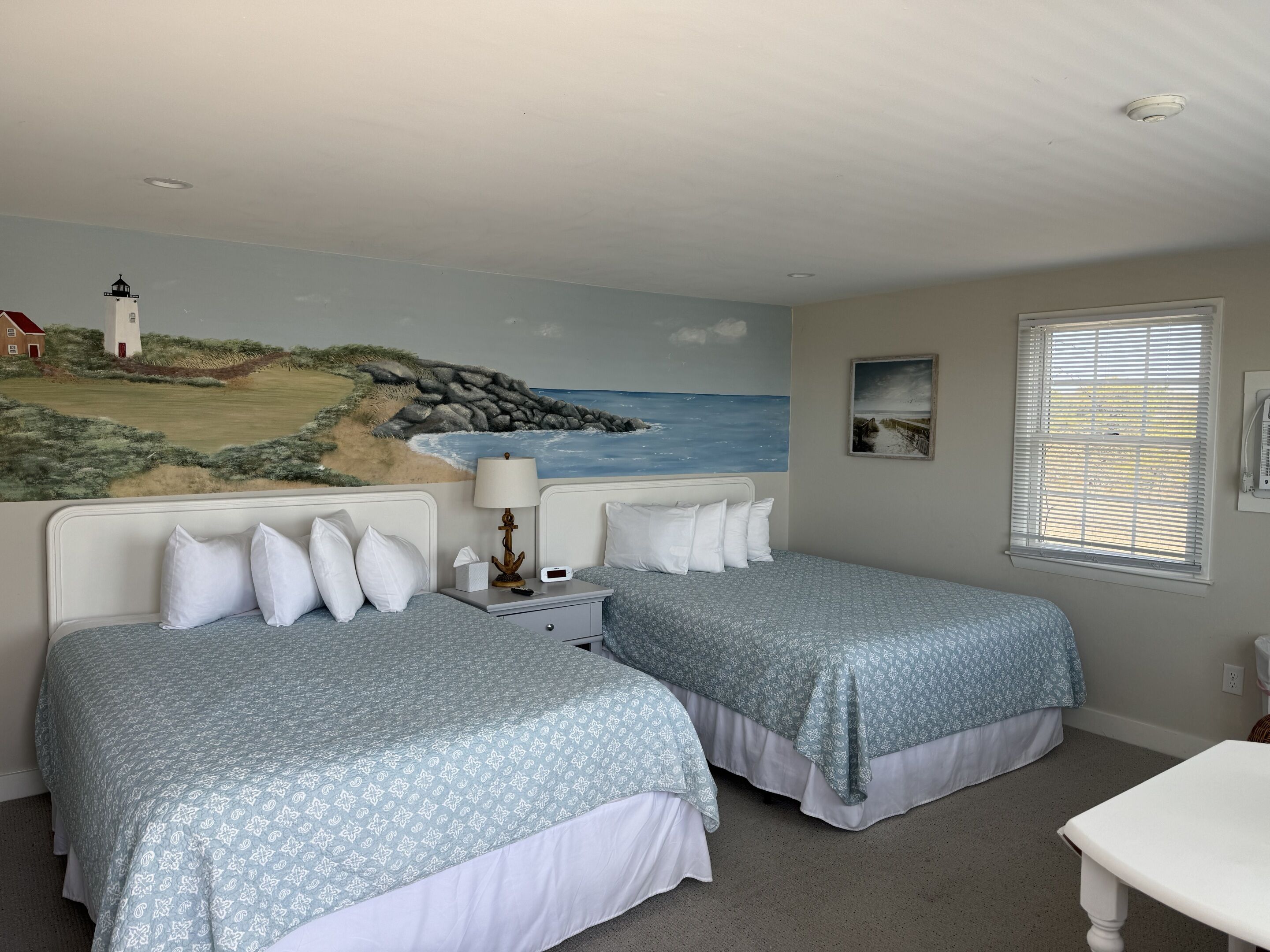 A bedroom with two beds, a nightstand with a lamp, a painting of a coastal scene, a window with blinds, and a small bedside table.