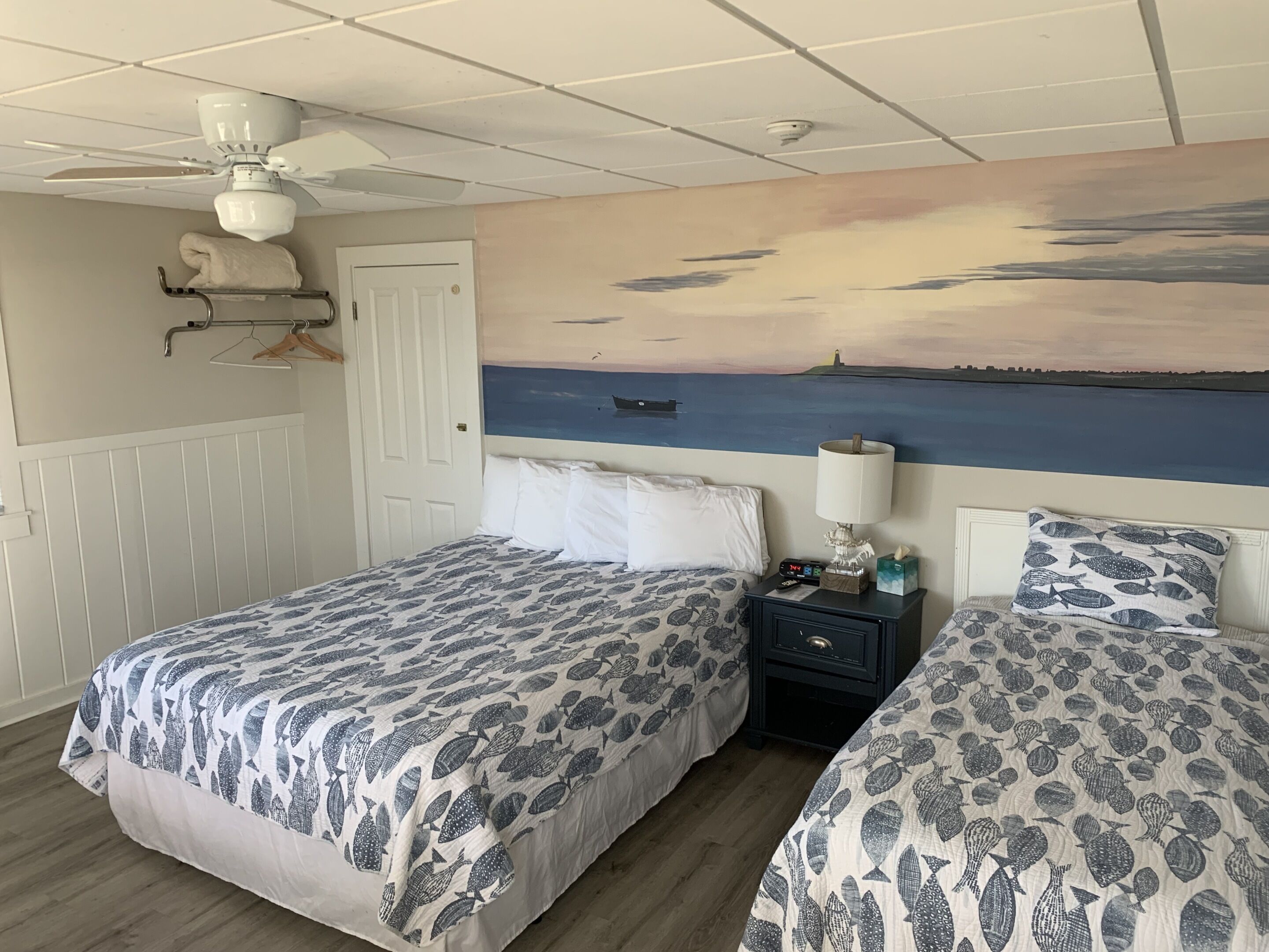A bedroom with two beds, a ceiling fan, a nightstand with a lamp, a painting of a seascape, and a door.
