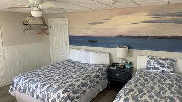 A bedroom with two beds, a ceiling fan, a nightstand with a lamp, a painting of a seascape, and a door.