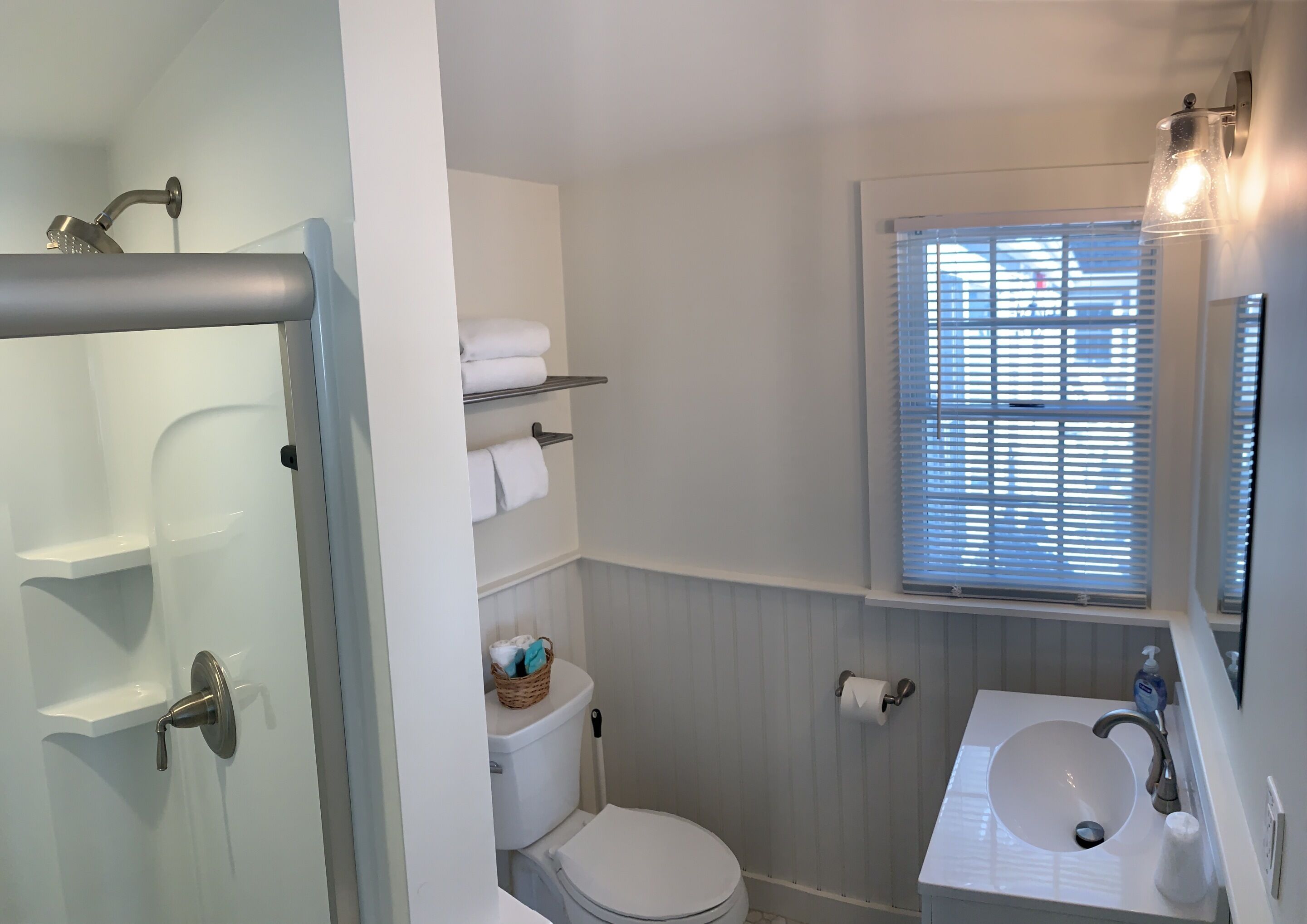 A bathroom with a toilet, a sink, a window with blinds, and a shower area.
