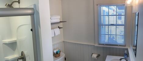 A bathroom with a toilet, a sink, a window with blinds, and a shower area.