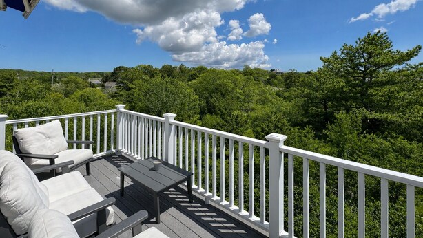 A deck with outdoor furniture, a white railing, and a view of a forested landscape. - The Ocean View (Centerville)
