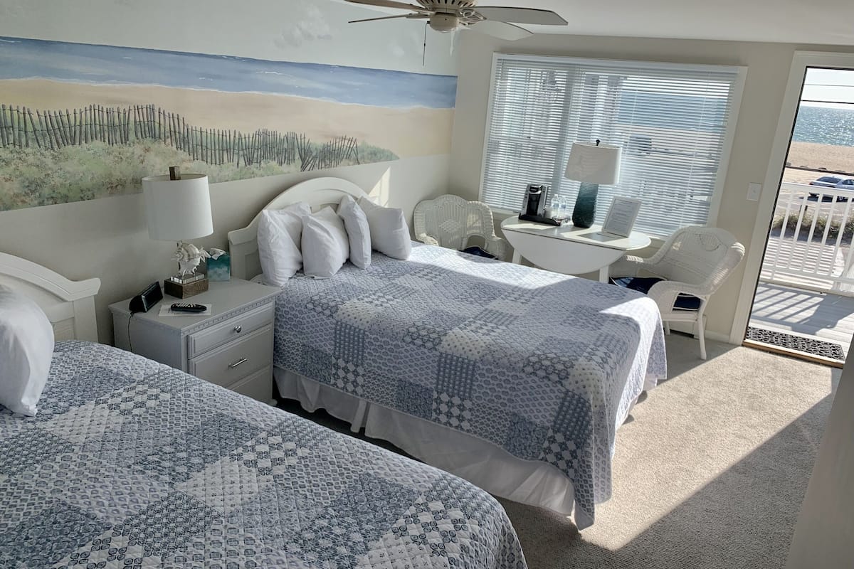 A hotel room with two beds, a ceiling fan, a beach mural, a window with blinds, a small table with a lamp, and a chair.