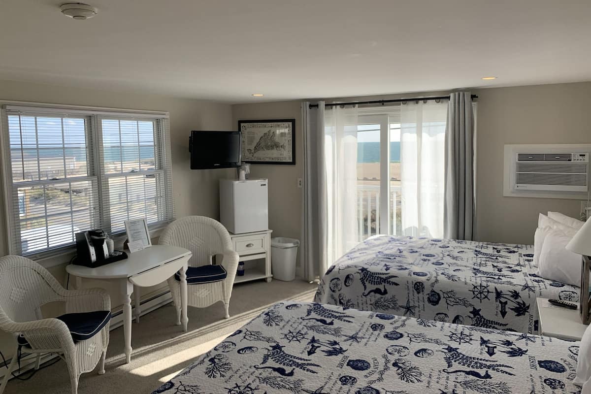 A hotel room with two beds, a desk, a chair, a television, and a window with a view of the beach.