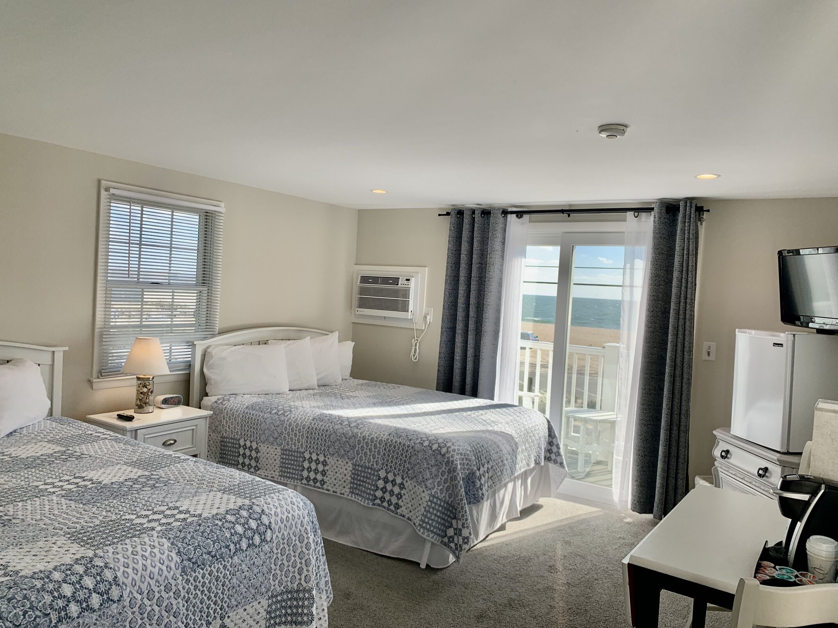 A hotel room with two beds, a TV, a refrigerator, and a balcony with a view of the beach.