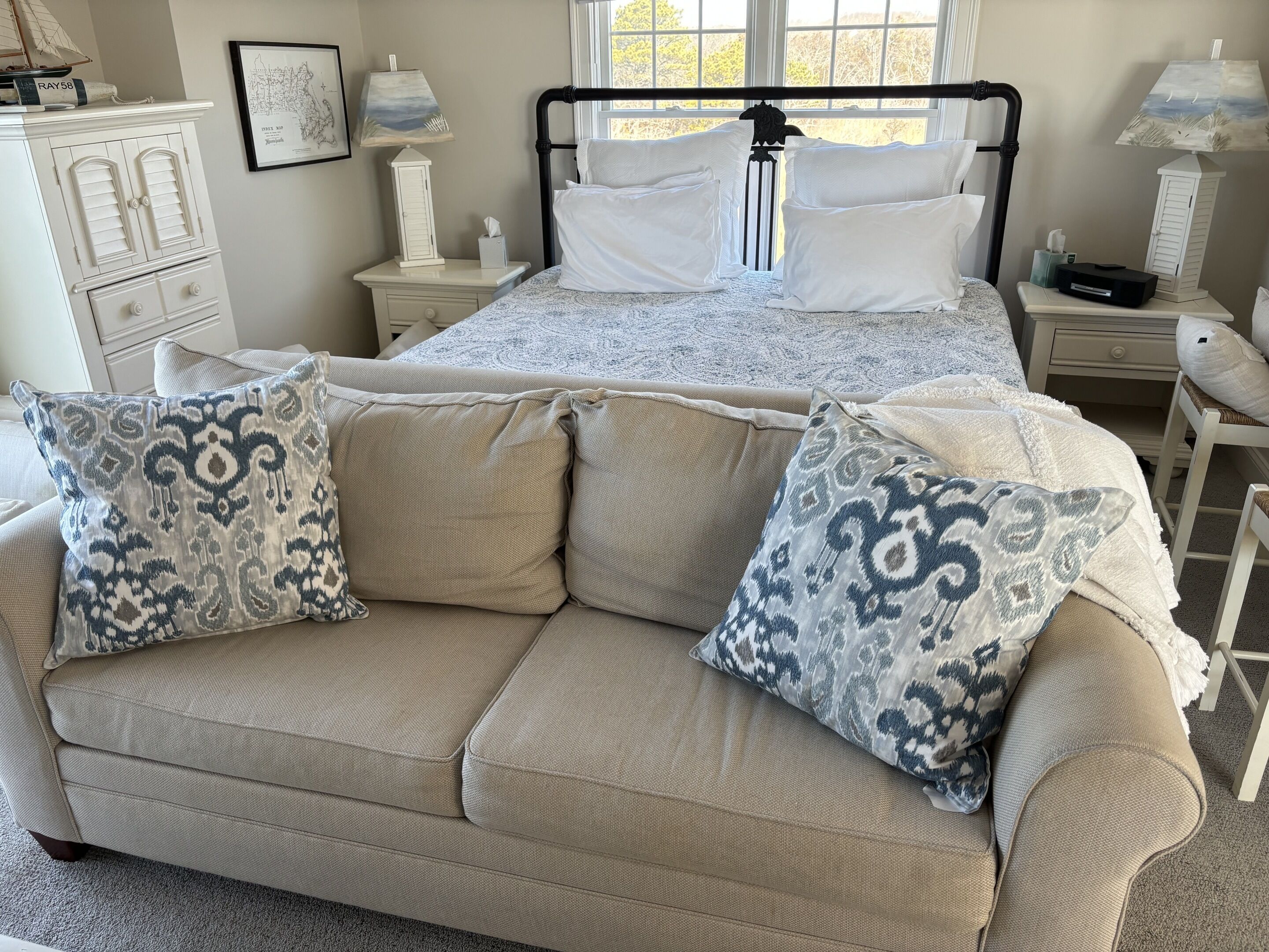 Een slaapkamer met een bed, een beige bank, gestreepte kussens, een nachtkastje, een dressoir en een raam met uitzicht.