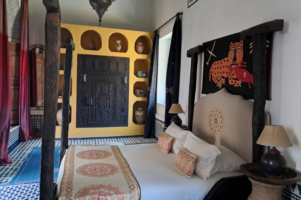 Une pièce comprenant un lit, une étagère fixée au mur ornée d’objets décoratifs, une grande porte aux motifs complexes et une lampe suspendue.