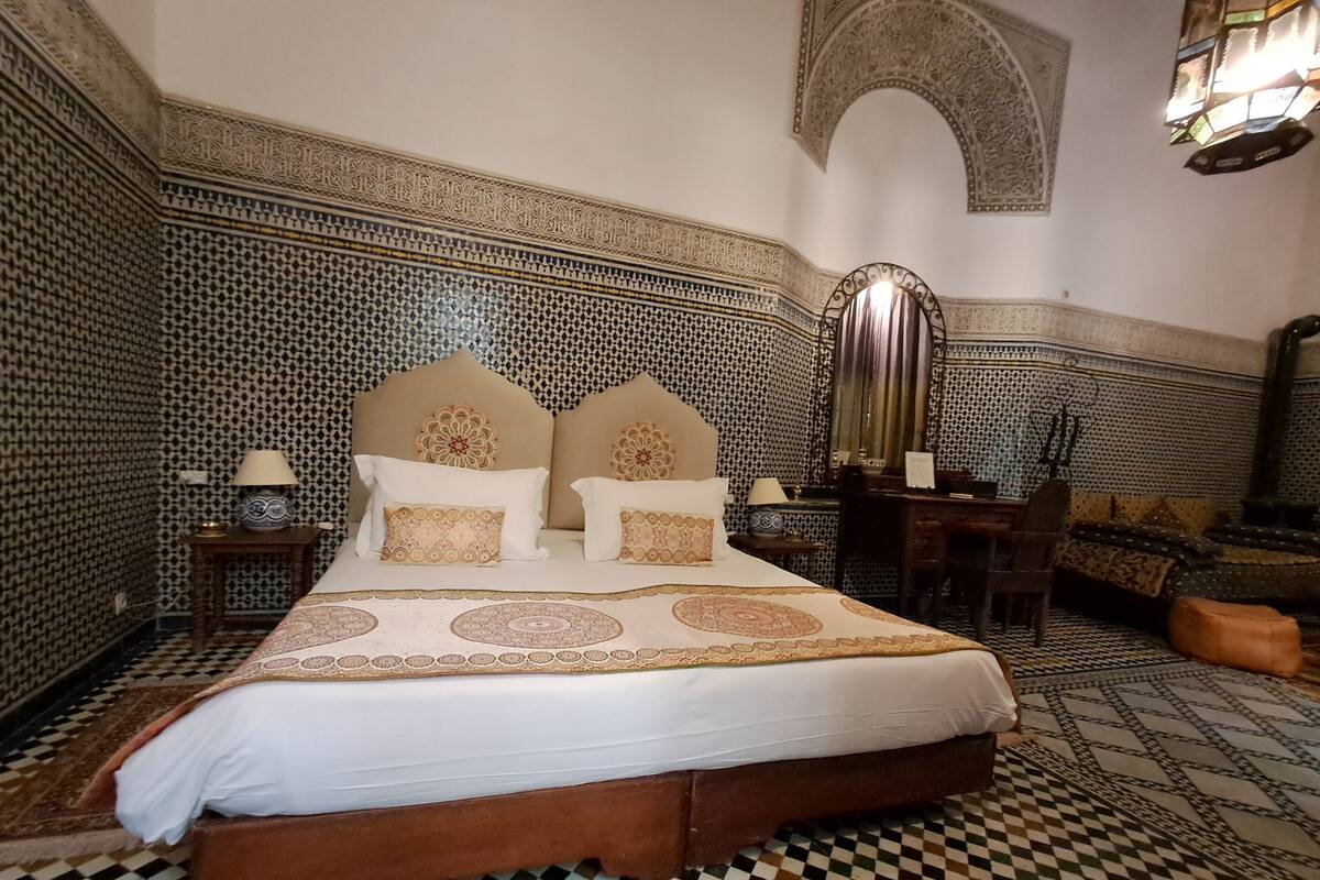 Un lit avec une couvre-lit à motifs, une tête de lit ornée d’éléments décoratifs et un sol à carreaux.
