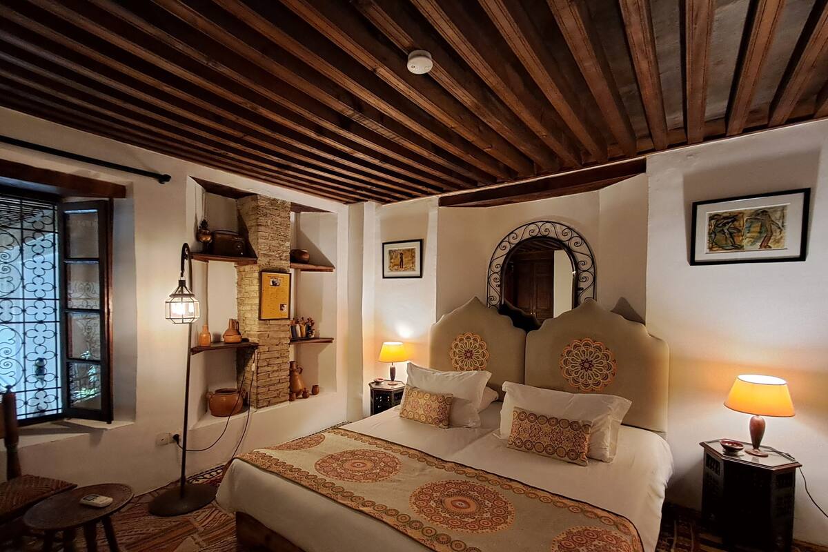 Une chambre confortable avec un plafond en bois, un grand lit recouvert d’une literie à motifs, un mur en pierre et une petite table avec une lampe.
