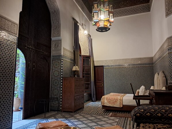 Une pièce traditionnelle marocaine avec un sol carrelé aux motifs géométriques, une porte en bois, un lit recouvert d’une courtepointe à motifs et une lanterne suspendue.