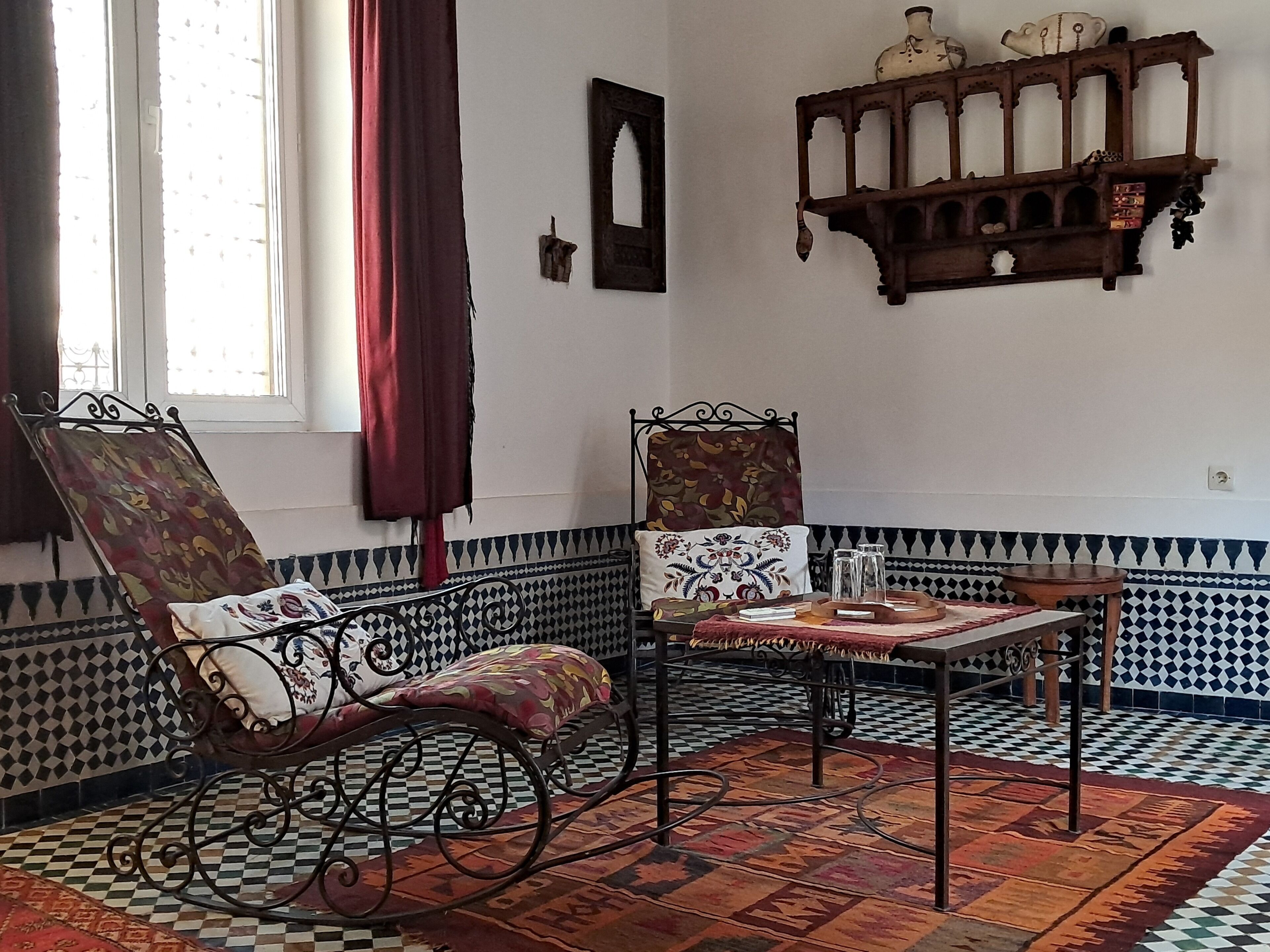 Um quarto com piso xadrez, uma cadeira estampada, uma mesa com bandeja e uma prateleira na parede com objetos decorativos.