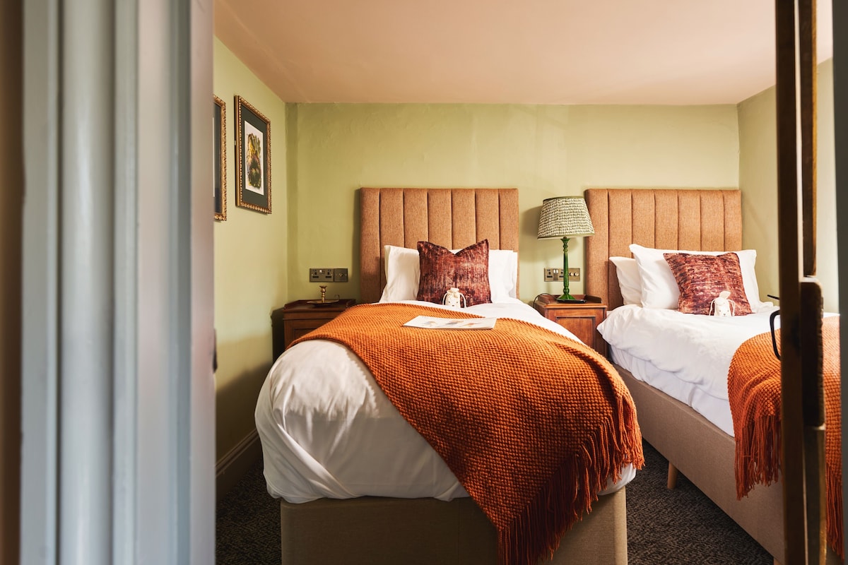 Habitación de hotel con dos camas, cada una con cabecera, mesita de noche, lámpara y un cuadro en la pared.