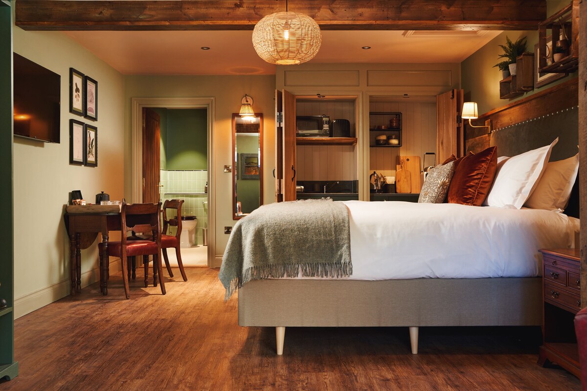 A bedroom with a bed, a desk, and a chair. There is a door leading to a bathroom and a kitchen.
