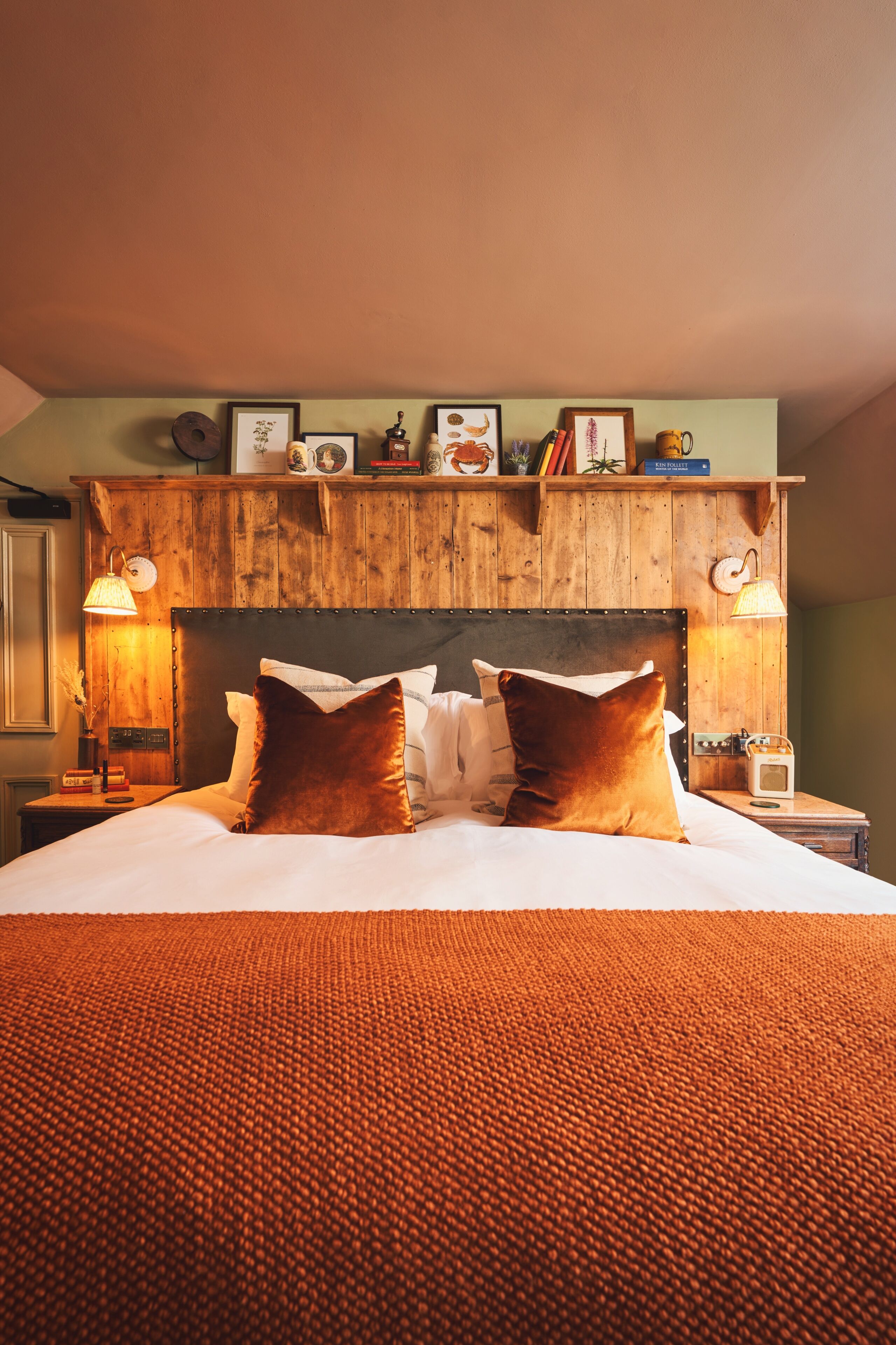 A hotel room with a wooden headboard, a bed with two pillows, a bedside table with a lamp, and a shelf with various items.