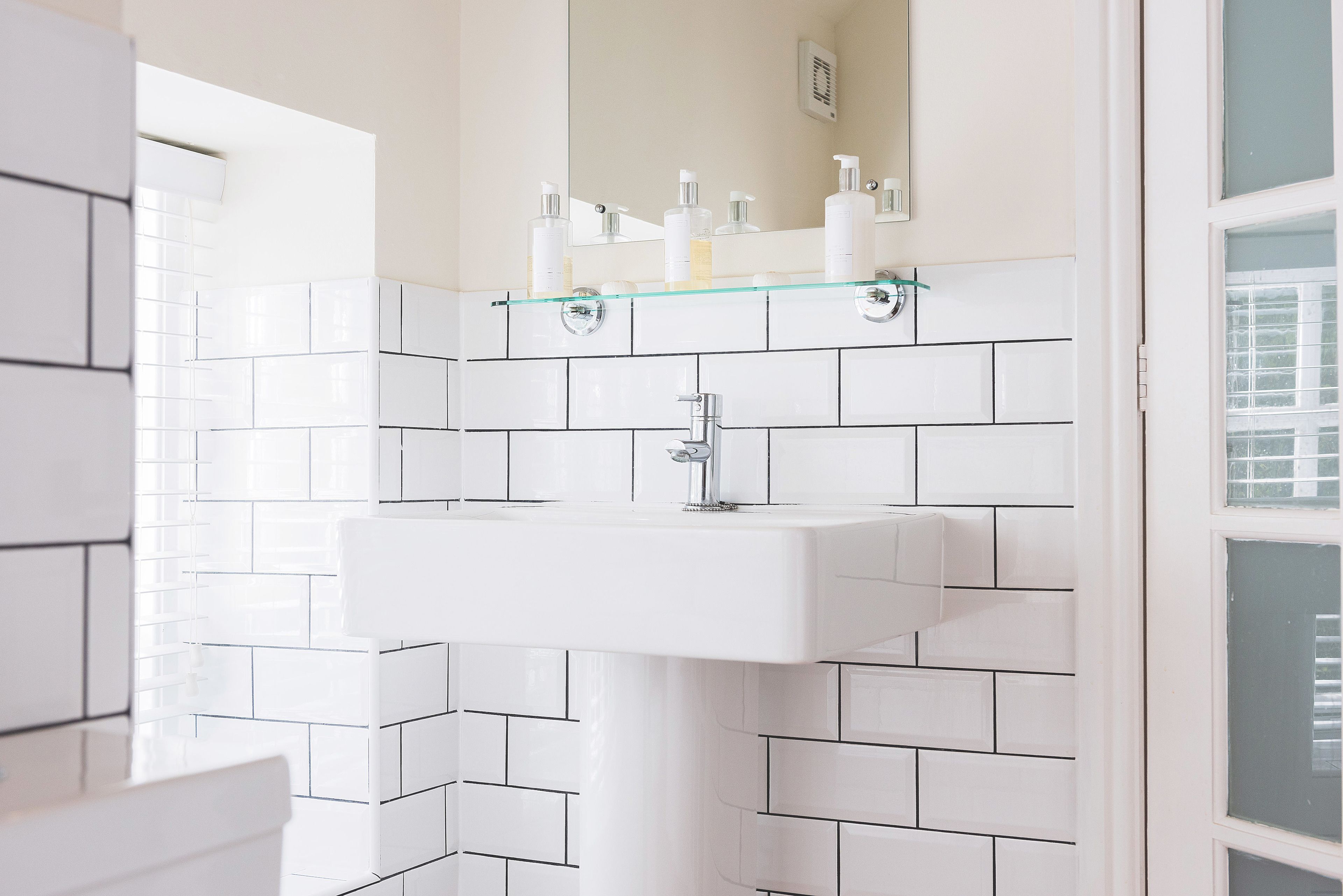 A bathroom with white tiled walls, a glass shelf with toiletries, and a white sink.