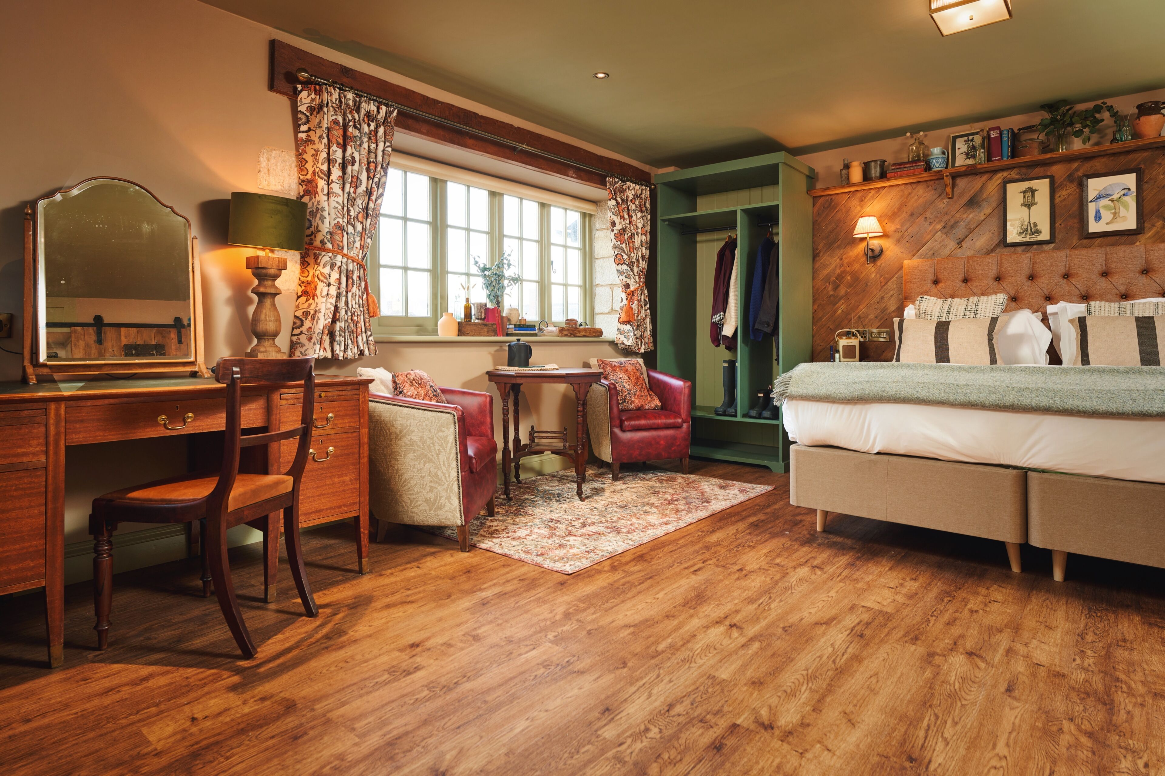 Una habitación tradicional con piso de madera, un escritorio con espejo, un sillón rojo, una mesita, un sofá y una cama.