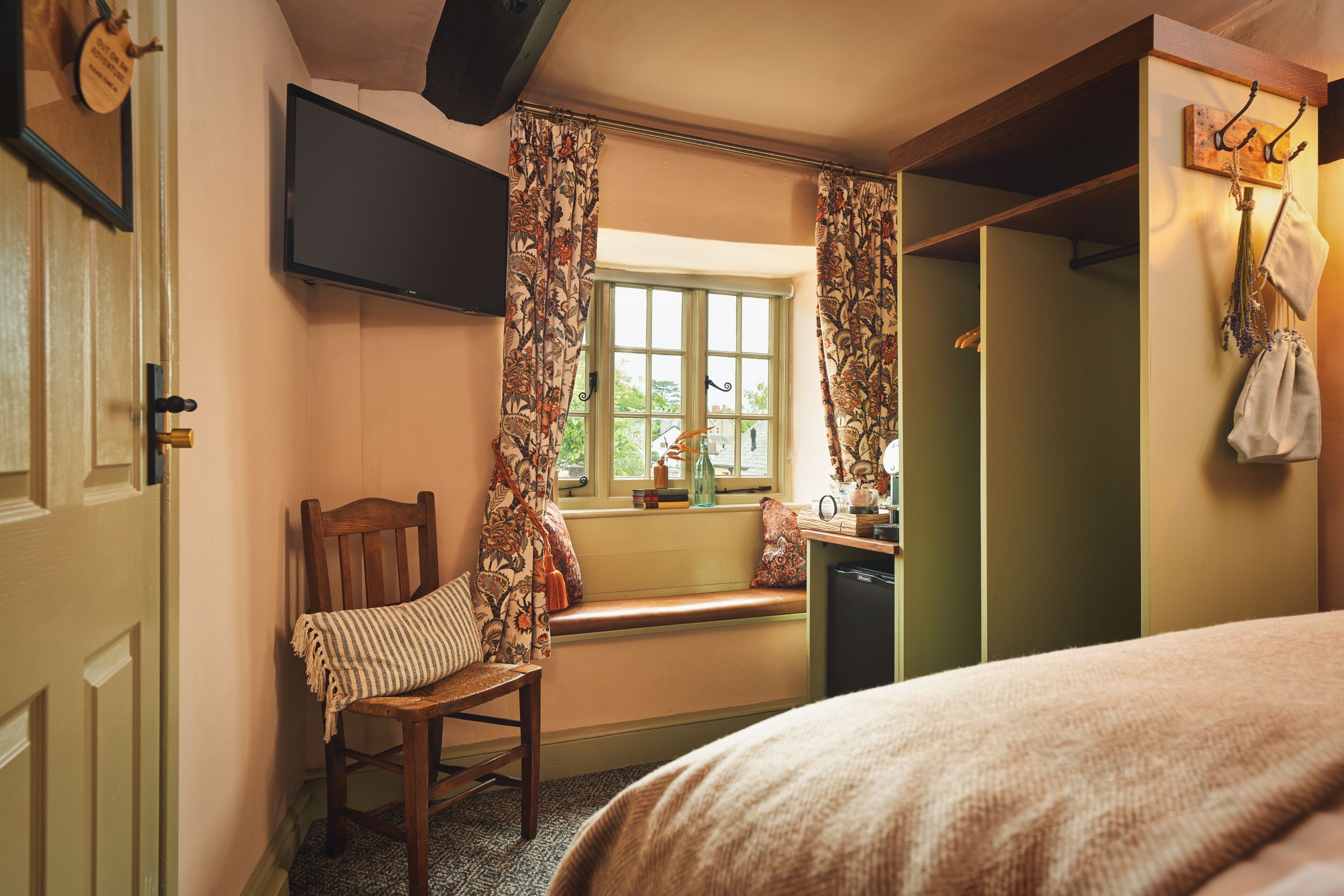 A room with a bed, a chair, a television, a window with curtains, a bench, and a small table with books and a vase.