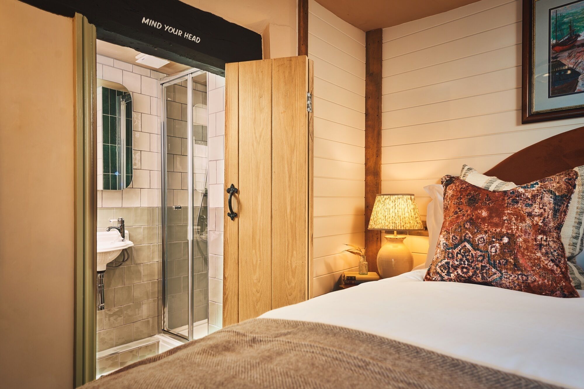 Habitación de hotel con cama, lámpara de noche, armario de madera y baño con cabina de ducha de vidrio.