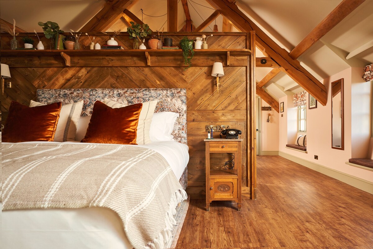 A bedroom with a wooden ceiling, a bed with white bedding, a nightstand with a lamp, and a window with a view of the outside.