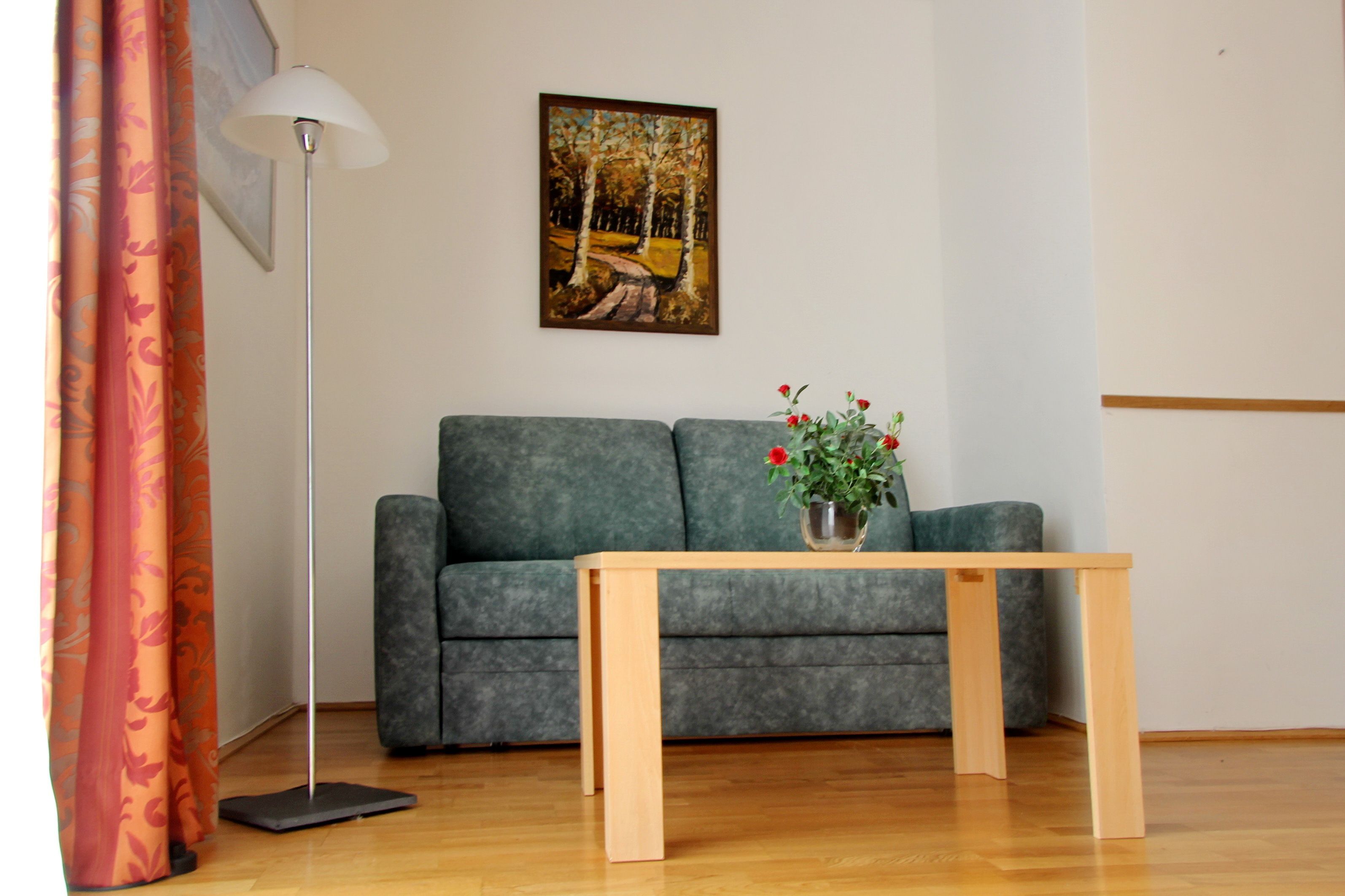 Un salon avec un canapé gris, une table basse en bois sur laquelle est posé un vase de fleurs rouges, un lampadaire et un tableau représentant une scène de forêt accroché au mur.