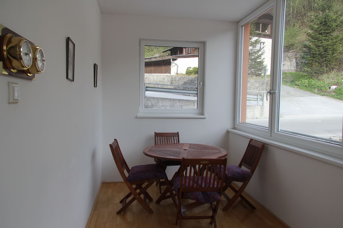 A dining area with a round table and four chairs, a window with a view of a building and greenery, and a wall-mounted clock.