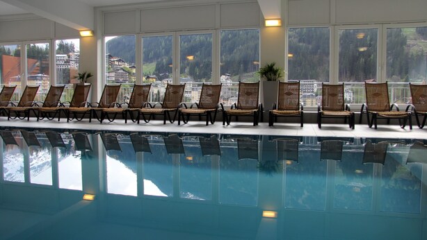 Ett område vid poolen med stolar som är vända mot poolen, stora fönster med utsikt över ett bergigt landskap och ett kristallklart poolvatten.