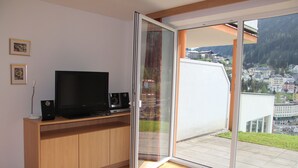 Ett modernt vardagsrum med en platt-TV placerad på ett träskåp, och en skjutdörr i glas som leder ut till en balkong med utsikt över staden.