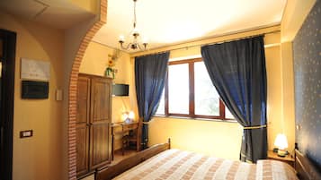 A bedroom with a bed, a desk, and a chair. There is a window with curtains, a chandelier, and a wall-mounted TV.