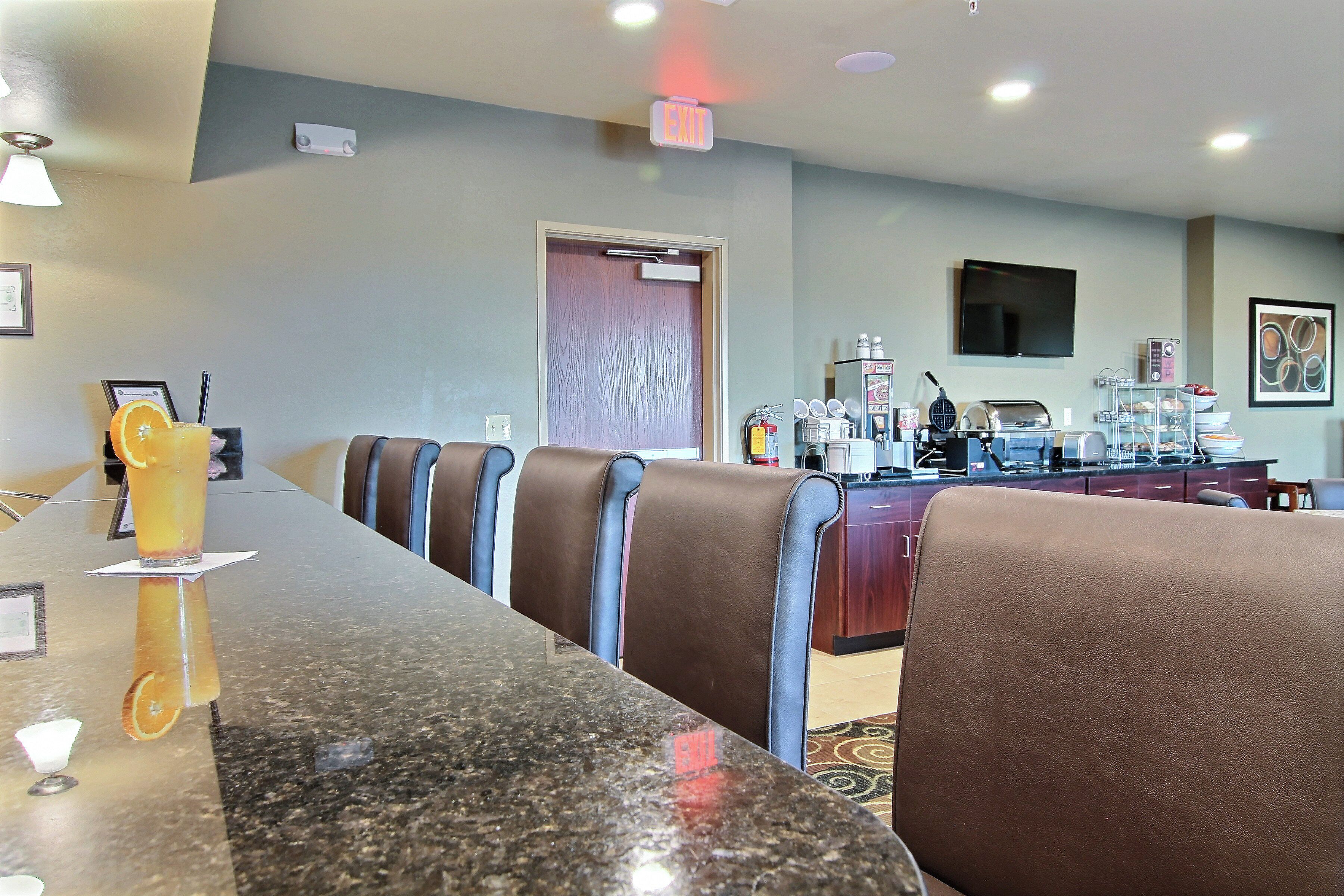 A hotel dining area with a long table, brown cushioned chairs, a bar area with various items, and a television on the wall. - Cobblestone Hotel & Suites - Beulah (Beulah)
