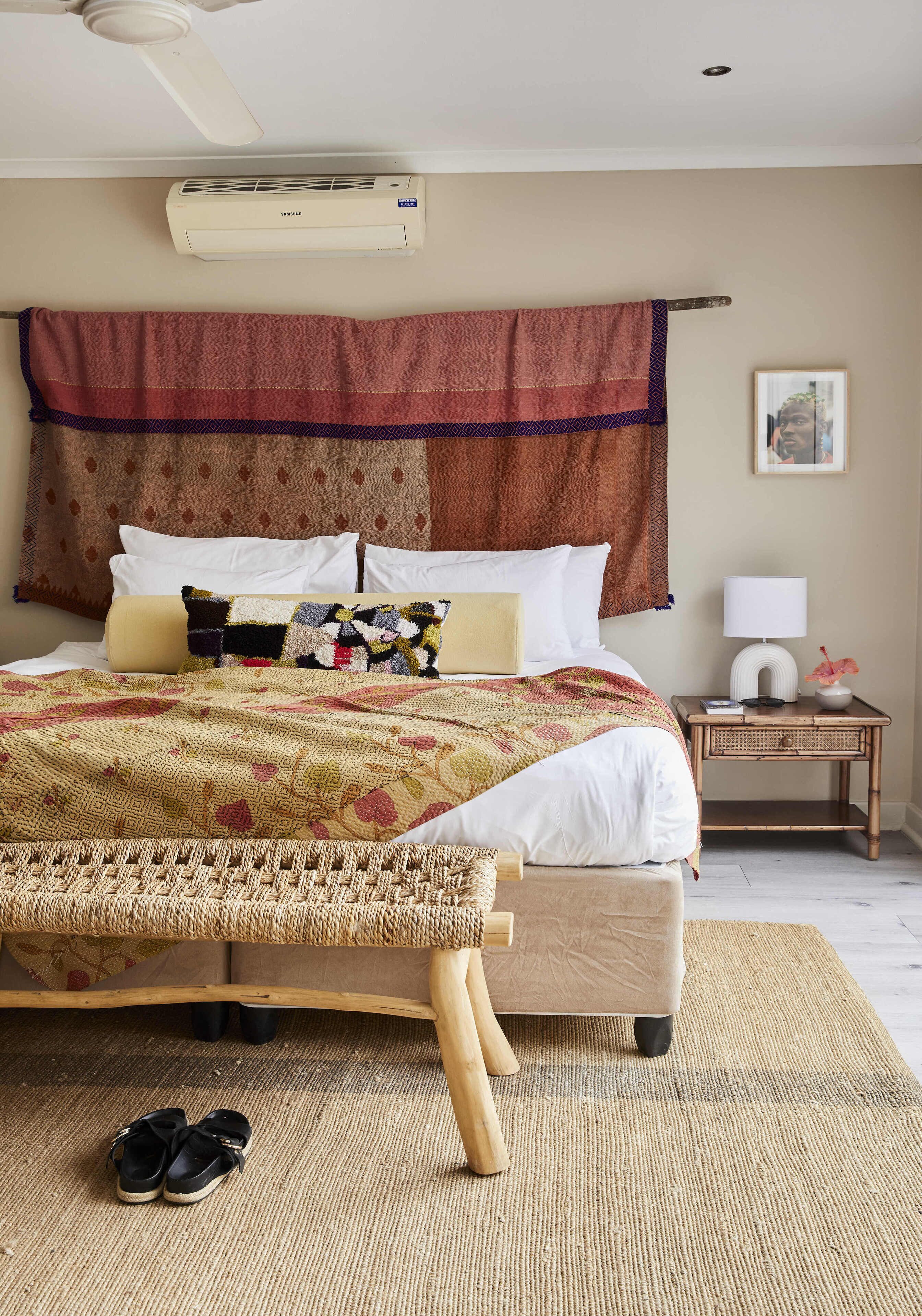 Uma cama bem arrumada com um cobertor estampado, uma mesinha de cabeceira com uma luminária e uma pequena planta, e um ar-condicionado fixado na parede.